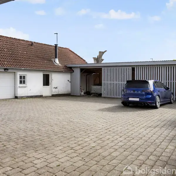 En hvid bygning med rødt tegltag, tilstødende en garage. En blå bil holder parkeret foran på en gårdsplads med stenbelægning. Himlen er delvist skyet.