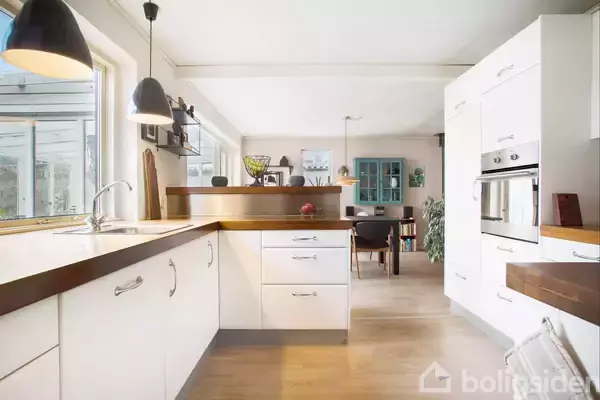 Et moderne køkken med hvide skabe, træbordplader og indbyggede ovne. Køkkenet åbner ud til en spisestue med et bord og stole. Naturligt lys strømmer ind fra vinduerne.