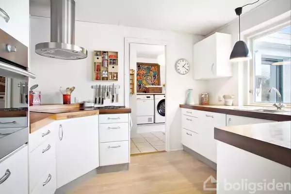 Et hvidt, moderne køkken med træborde og en kogeø. Der hænger en emhætte over øen, og et analogt ur ses på væggen. I baggrunden er en vaskemaskine i en tilstødende vaskerum.