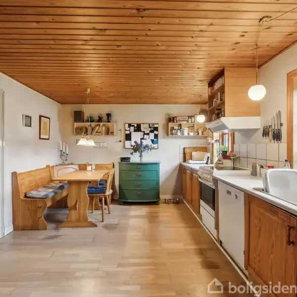 Køkken med træloft og trægulv. Køkkenbord med vask langs vinduer. Spiseplads med bænk i hjørne. Grøn kommode, hylder, opslagstavle på væggen. Behagelig og lys atmosfære.