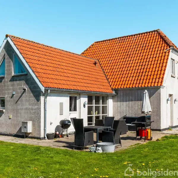 Et hus med røde tegltage og grå vægge står i en have. Havemøbler og en grill er placeret på terrassen foran huset under en klar blå himmel.