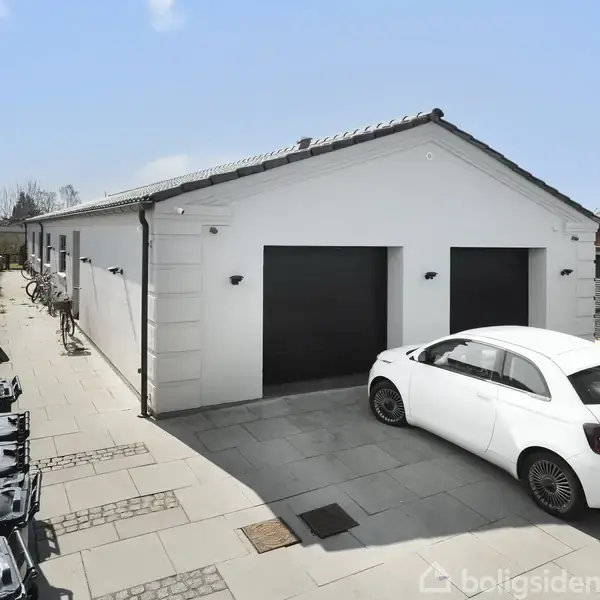 En hvid bil holder parkeret foran en moderne, hvid garagebygning med to sorte porte. Omgivet af en flisebelagt gårdsplads med affaldscontainere og cykler langs væggen.