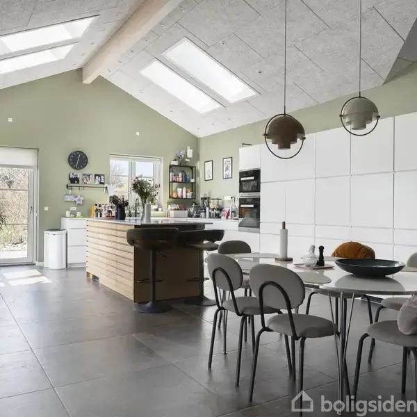 Et moderne køkken med åbent loft og ovenlysvinduer. Køkkenøen har barstole, det runde spisebord har stole omkring, og hvide skabe dækker bagvæggen. Planter pynter rummet, der har en grøn nuance.