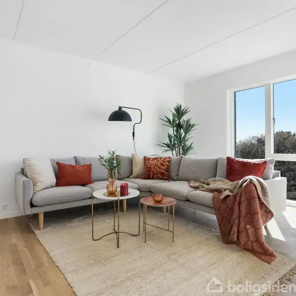 Grå sofa med røde puder står i hjørnet af en lys stue. To runde borde med en grøn plante dekorerer rummet. Store vinduer giver udsigt til blå himmel.