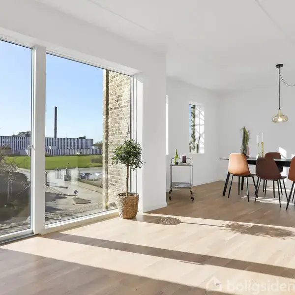 Spisestue med spisebord og stole i moderne stil. Sollys strømmer ind gennem store vinduer med udsigt til en grøn have og et industrielt område i baggrunden.