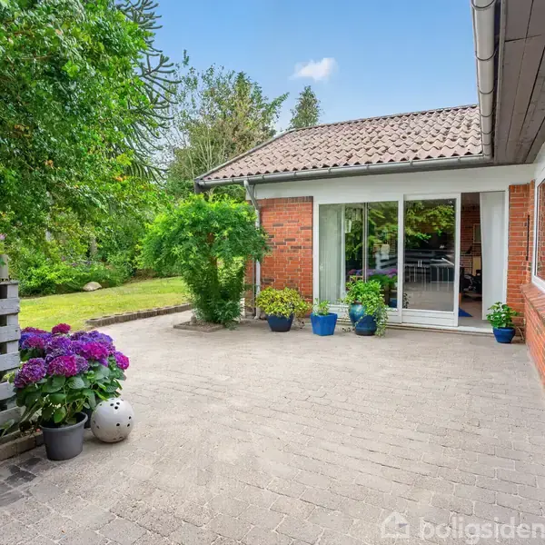 Terrasse med blomsterkrukker står foran et hus med rød murstensfacade og store glasdøre. Omgivet af grønne træer og buske i en have.