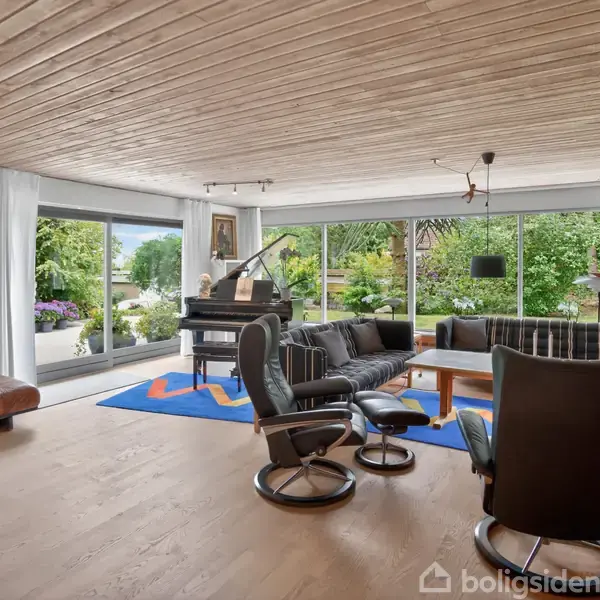 Stue med moderne møblering. En sort lænestol og sofa står ved et sofabord, under en stor træring. Et flygel står ved glasvinduer, der åbner ud til en frodig have. Ingen tekst.