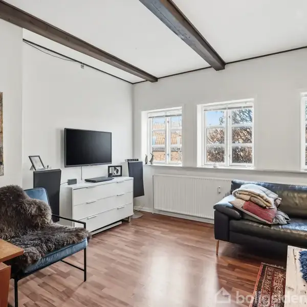 En stue med en sort lædersofa og et sofabord, placeret foran et vindue med tre rammer. Ved siden af sofaen står et hvidt skab med fjernsyn ovenpå.