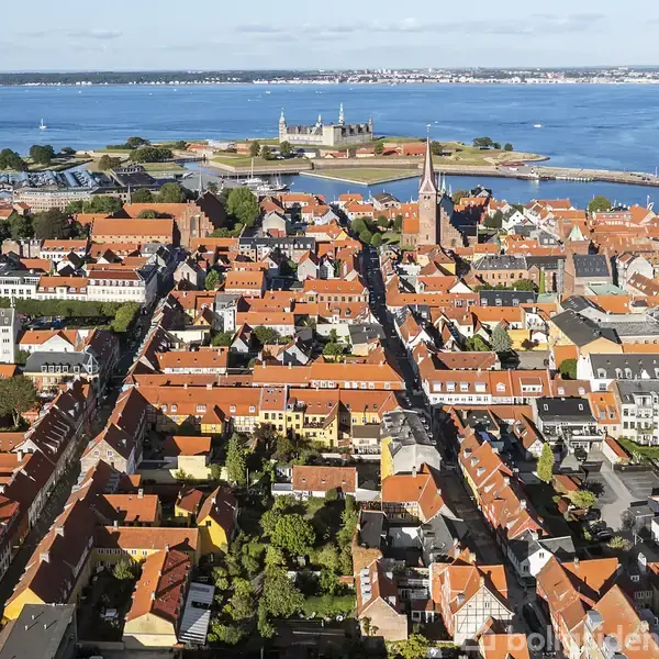 Bylandskab med tætte, røde tegltage, centralt kirketårn. Baggrund prydes af vandudsigt og en ø med et historisk slot. Klar blå himmel møder horisonten over havet.