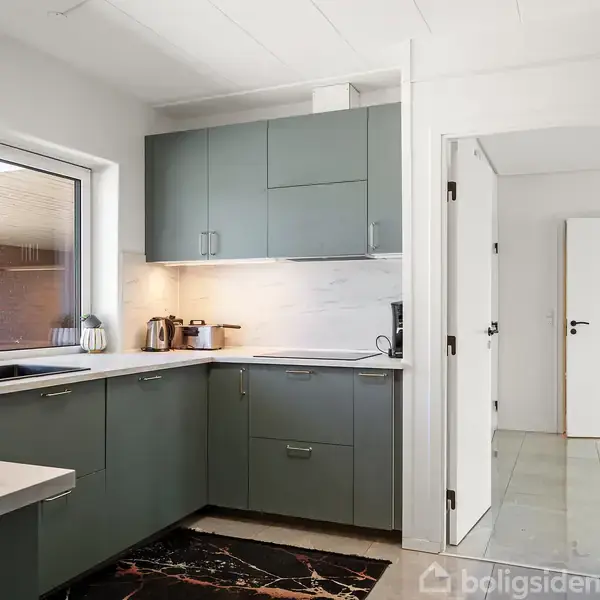 Køkken med grønne skabe, marmorflisegulv; ses i en moderne boligindretning med marmorbordplade, køkkenredskaber og en åbning mod en gang. Stort vindue med udsigt til murstensvæg.