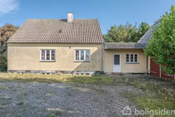 Et gult hus med rødt tegltag og to vinduer foran. Huset står ved en grusbelagt plads omgivet af træer og buske i baggrunden.