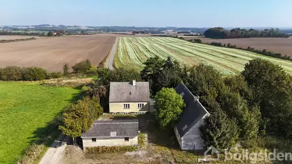 En gård med tre bygninger omgivet af træer og marker, der strækker sig ud i horisonten under en klar blå himmel.