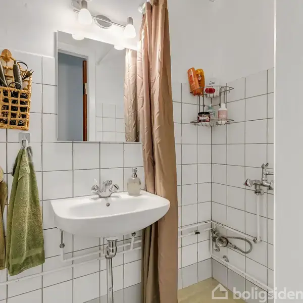 En hvid håndvask med spejl hænger på flisevæggen. Til venstre hænger håndklæder, og til højre er der en bruseniche med en hylde med forskellige produkter.