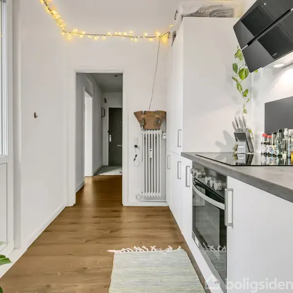 Et moderne køkken med hvidt inventar og sorte bordplader. Køkkenet er pyntet med lyskæder og planter. Der er en ovn og emhætte. En gang fører mod en dør i baggrunden.