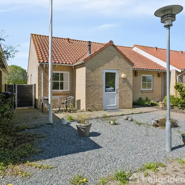Et lille hus med rødt tegltag står i en forhave med grusbelægning og spredte planter. Til venstre ses et skur og et flagstang. Solrig og klar himmel.