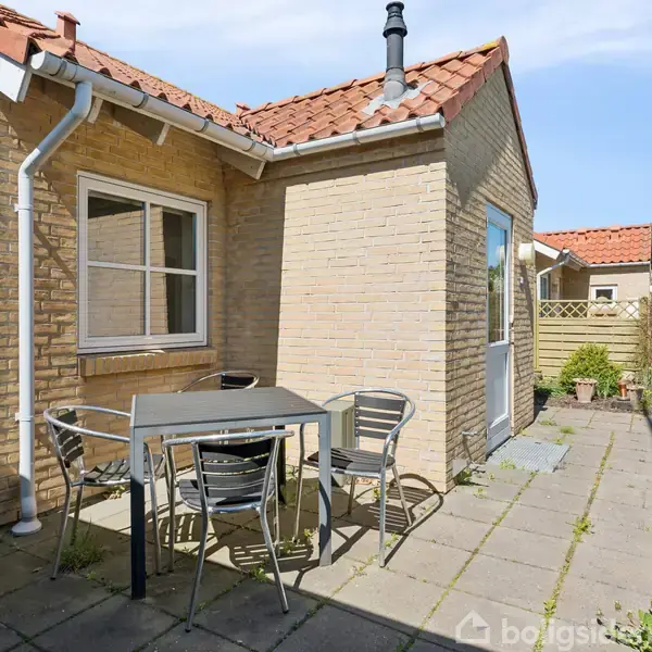 Flisebelagt terrasse med firkantet bord og fire stole står ved gulstenshus. Omkringliggende have med buske og græs under blå himmel.