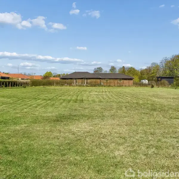 Græsplæne, nyklippet og grøn, strækker sig ud med huse og skure i baggrunden under en blå himmel med få skyer.