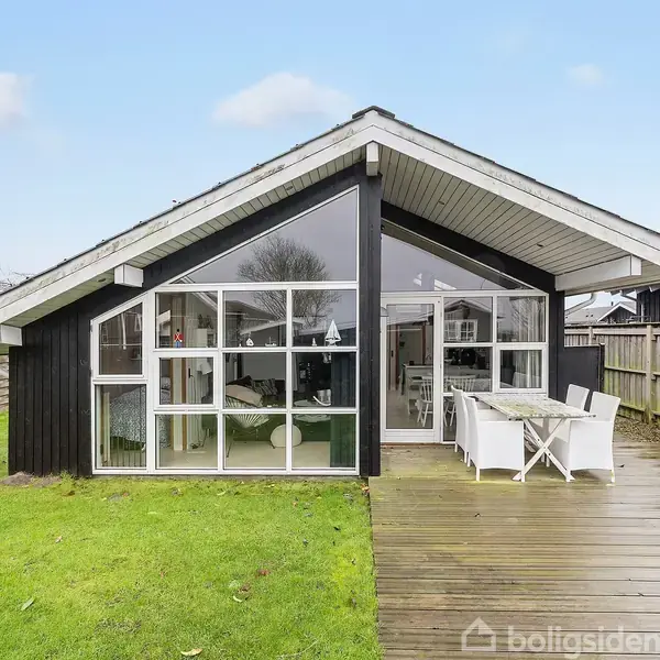 Sort træhus med store vinduer står stille. Terrasse med havemøbler og grill omkranser huset i en have med græsplæne. Stakit adskiller naboejendomme.