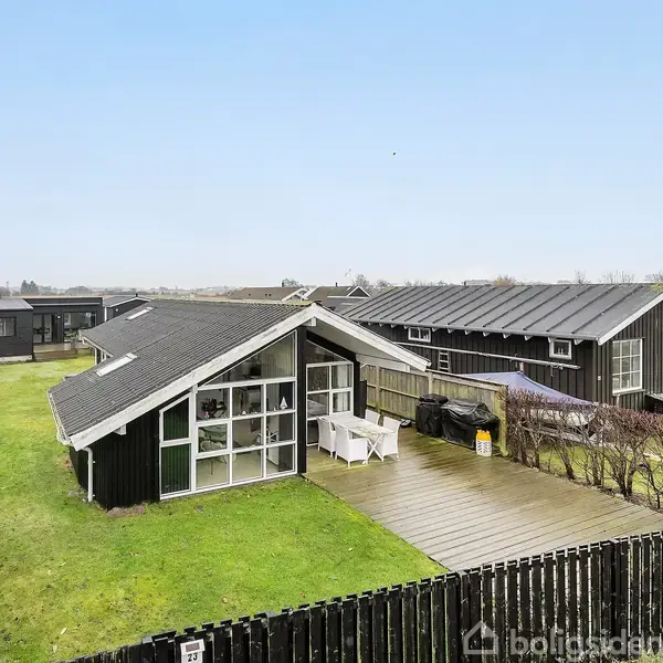 Sort sommerhus med store vinduer står på en velholdt græsplæne, omgivet af et trædæk med hvide havemøbler. Andre lignende bygninger ses i baggrunden under en klar himmel.