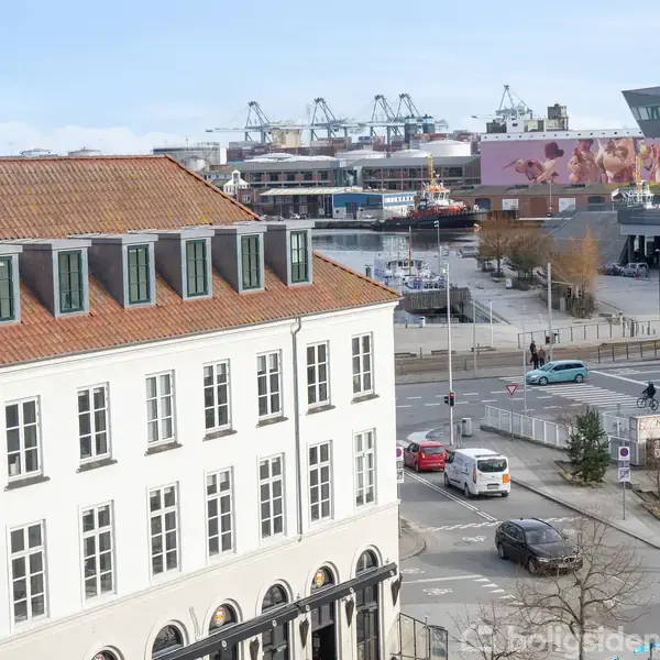 Et hvidt bygning med rødt tag står ved en vej med biler og cyklister. I baggrunden er der industrielt havneområde med kraner og moderne bygninger.