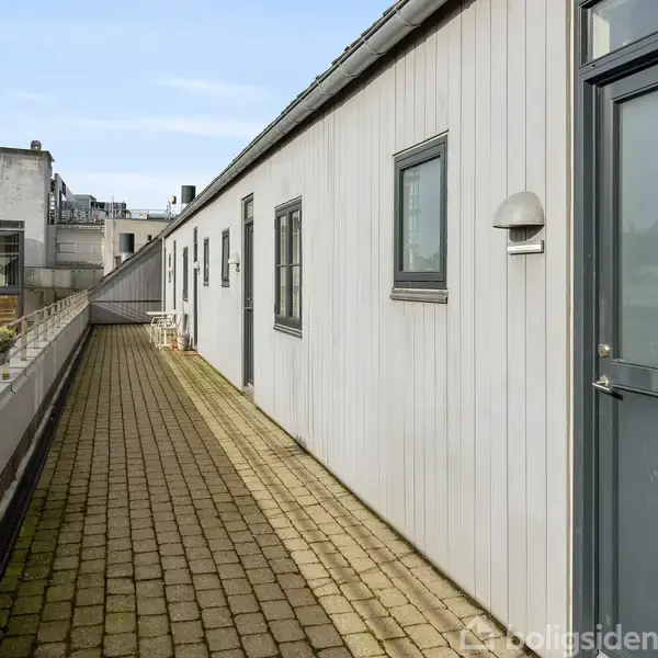 Dør og vinduer på en lang, hvid bygning flankerer en brolagt terrasse. Bygningen ligger i en bymæssig kontekst med stillads og omkringliggende bygninger set i baggrunden.