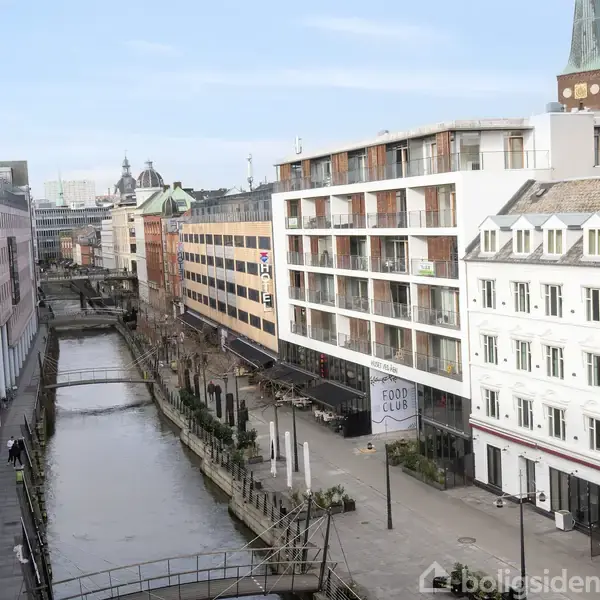 En række moderne bygninger står langs en kanal. En gågade løber parallelt med vandet. Tegnet "FOOD CLUB" hænger på en af bygningerne. Himlen er klar, og dagtimerne er lyst op.