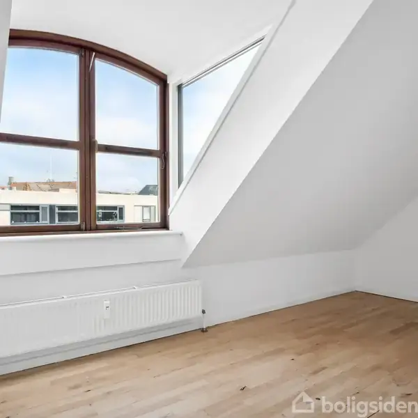 Et tomt værelse har et trægulv og et stort, buet vindue med udsigt til bygninger udenfor. Rummet har skråvægge og en radiator under vinduet.