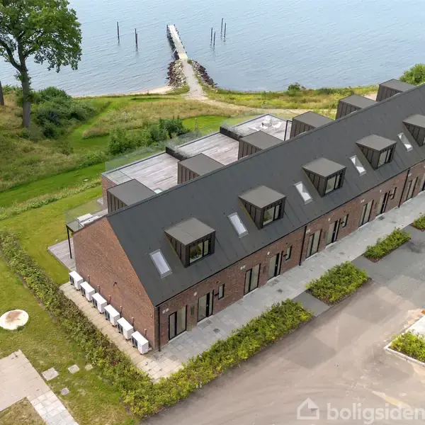 Terrasse med to polstrede stole står stille med udsigt over en stor sø. Murstensvægge omkranser området, mens store glaspartier giver udsigt til en klar blå himmel med spredte skyer.