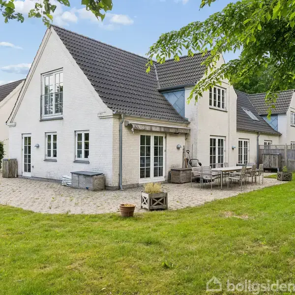 Hvidt hus med mørkt tag står stille på en flisebelagt terrasse. Havemøbler står ved siden af, omgivet af grøn græsplæne og træer.