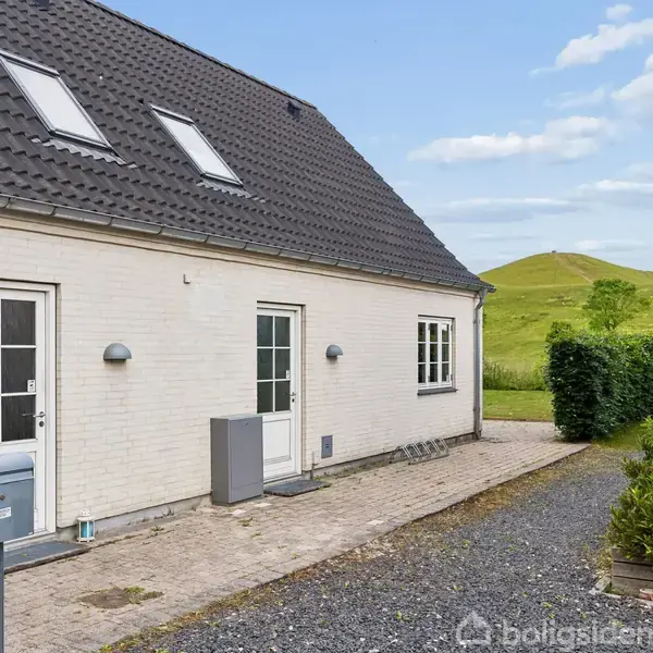 En hvid murstensbygning med sort skråt tag står stille på en grusvej. Omkring bygningen er der grønne buske og et baggrundslandskab med frodige bakker under en blå himmel.
