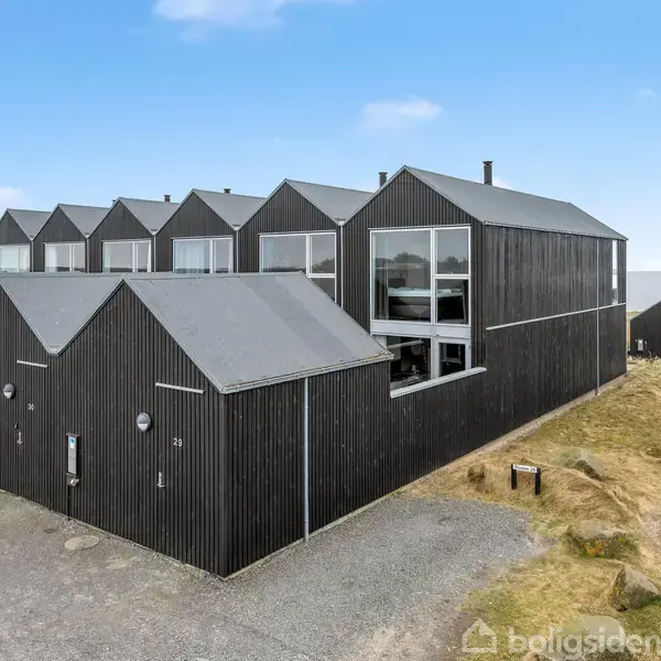 Sort hus med skarpt skrånende tag står stille på en grusvej. Omkring det ligger græsklædte klitter ved kysten.