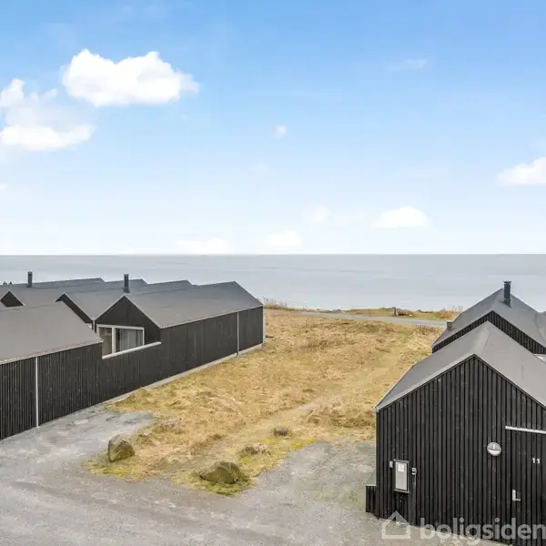 Sortmalede huse står stille ved en kyststrækning. Husene har moderne design og ligger tæt på en grå vej. Baggrund viser havet under en let skyet himmel.
