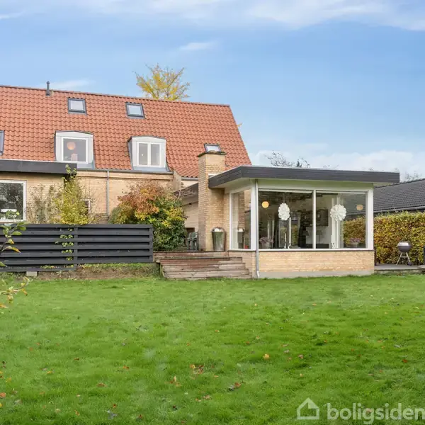 Et to-etagers hus med rødtag og store vinduer står i en grøn have med græsplæne. En terrasse med glasparti til venstre. Omkranset af buske og træer.