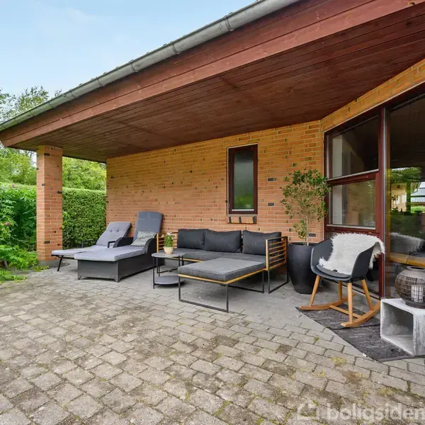 Terrassemøbler står på en flisebelagt terrasse ved et gult murstenshus med overdækning. Baggrunden viser grønne buske og træer, der giver en rolig, naturlig atmosfære.