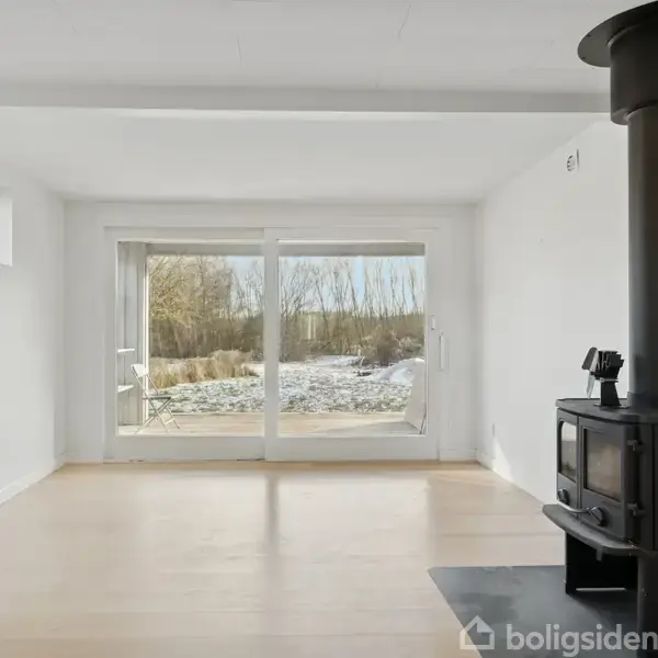 En sort brændeovn står i et hvidt rum med trægulv, placeret ved en glasdør, der åbner ud mod en sneklædt have.