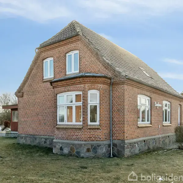 Rødt murstenshus står på en grund med græsplæne. Huset har flere buede vinduer og et skråt tag. I baggrunden ses en mindre bygning samt træer og himmel.