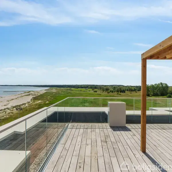 Træterrasse med glasrækværk står under en klar blå himmel ved kysten. En enkel pergola giver lidt skygge mens havet og en grøn skovkant ses i baggrunden.