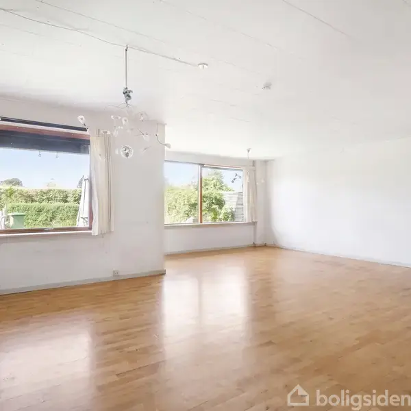 Stor stue med trægulv, sollys strømmende ind gennem store vinduer. Hvide vægge og loft. Omkringliggende grønt område ses udenfor gennem vinduerne.