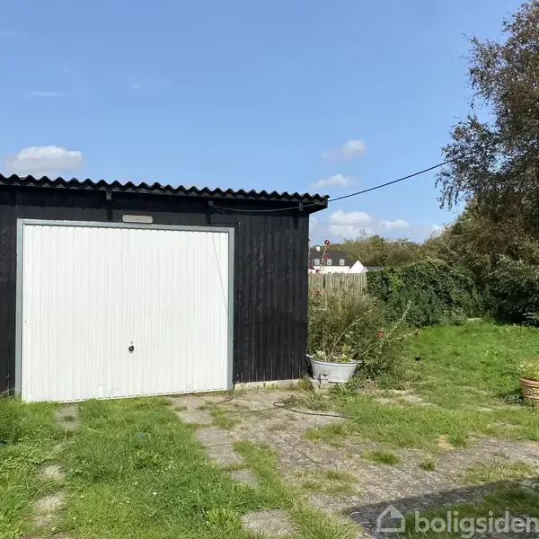 Et sort skur med en hvid dør står på en græsplæne omgivet af planter og træer, under en klar blå himmel. En sti fører op til døren.