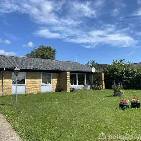 Et enfamiliehus i gule mursten står på en grøn græsplæne. Foran huset er der blomsterkrukker, og himlen er blå med skyer. En sti fører op til huset.