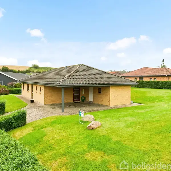 En etplans murstenshus med gråt tag står midt på en stor, velplejet græsplæne. Omkring huset er der hække og en stenlagt sti. Himmel er blå med få skyer.