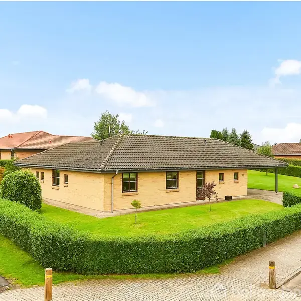 Et gult murstenshus står stille omgivet af en veltrimmet hæk i et roligt villakvarter med andre huse i baggrunden.