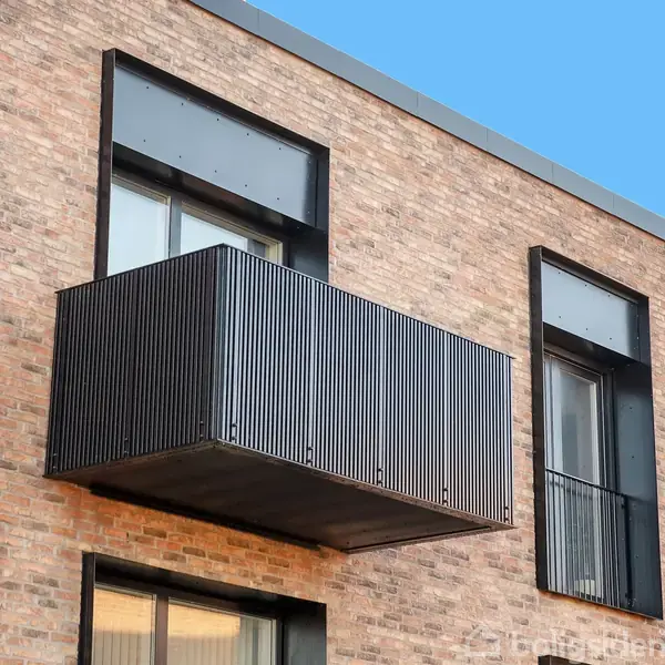 En balkon med en moderne metalfront hænger ud fra en murstensfacade. Omkringliggende vinduer er indrammet med mørke rammer. Baggrunden er en klar blå himmel.
