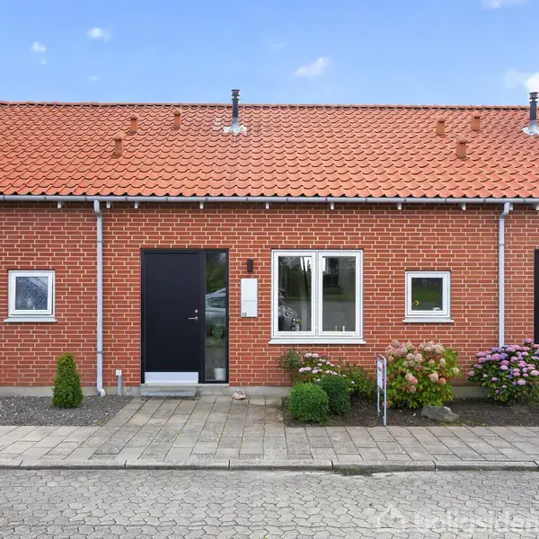 Rækkehus med rødt tegltag og facade i røde mursten, står stille, flankeret af vinduer og sorte døre. Foran ses fortov, små buske og blomster i en velholdt have.