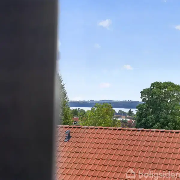 Et udsigt over et rødt tegltag ses gennem en åbning. Der er træer i forgrunden og en sø eller fjord i baggrunden under en blå himmel.