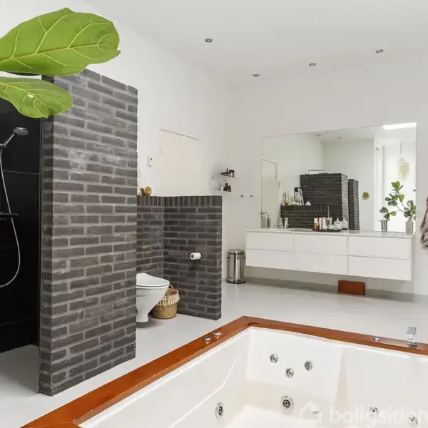 Jacuzzi omgivet af hvidt gulv i moderne badeværelse; bruseniche til venstre med sorte fliser, stor spejl over håndvask, og opbevaringsreol på højre side. Grønt planteblad i forgrunden.