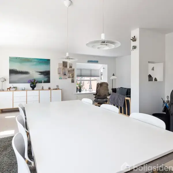 Spisebord med hvide stole står i et lyst rum. I baggrunden ses en brændeovn og et skab med kunst på væggen. Bordet er tomt, og omgivelserne er moderne.
