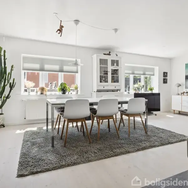 En spisestue med et stort bord og seks moderne stole placeret på et gråt tæppe. Rummet har store vinduer og er lyst med få dekorationer som planter og vægkunst.
