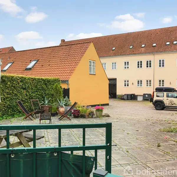 Gård med flisebelagt terrasse, hvor der står havemøbler og parasol. En gul bygning med tegltag omkranser gården. En bil er parkeret ved siden af affaldscontainere.
