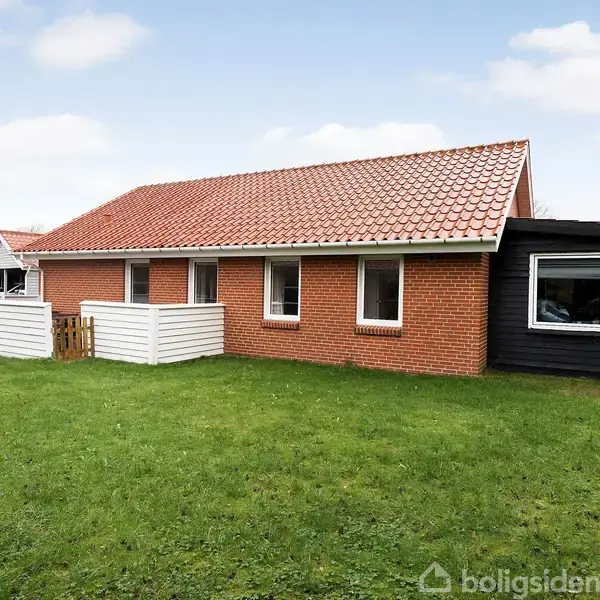 Rødt murstenshus med rødt tegltag, hvide vinduer og sort trætilbygning står på en græsplæne. Foran ses et hvidt stakit. Omkringliggende grønne områder giver et landligt præg.